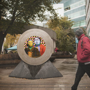 Masked person walks down the sidewalk on rainy day and passes oversized coin mounted on angular polished granite. The coin features life-size portraits of Chiefs Miskookenew and Kakekapenais in bright Pop Art colors, and features the text "Treaty No. 1", "1871", "3 dollars" and "Turtle Island".