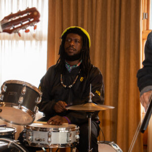 A musician wearing a black jacket and a bright green hat sits at a drumset playing the drums. In each corner of the image, there is a cut-off arm of the other musicians playing their instruments.