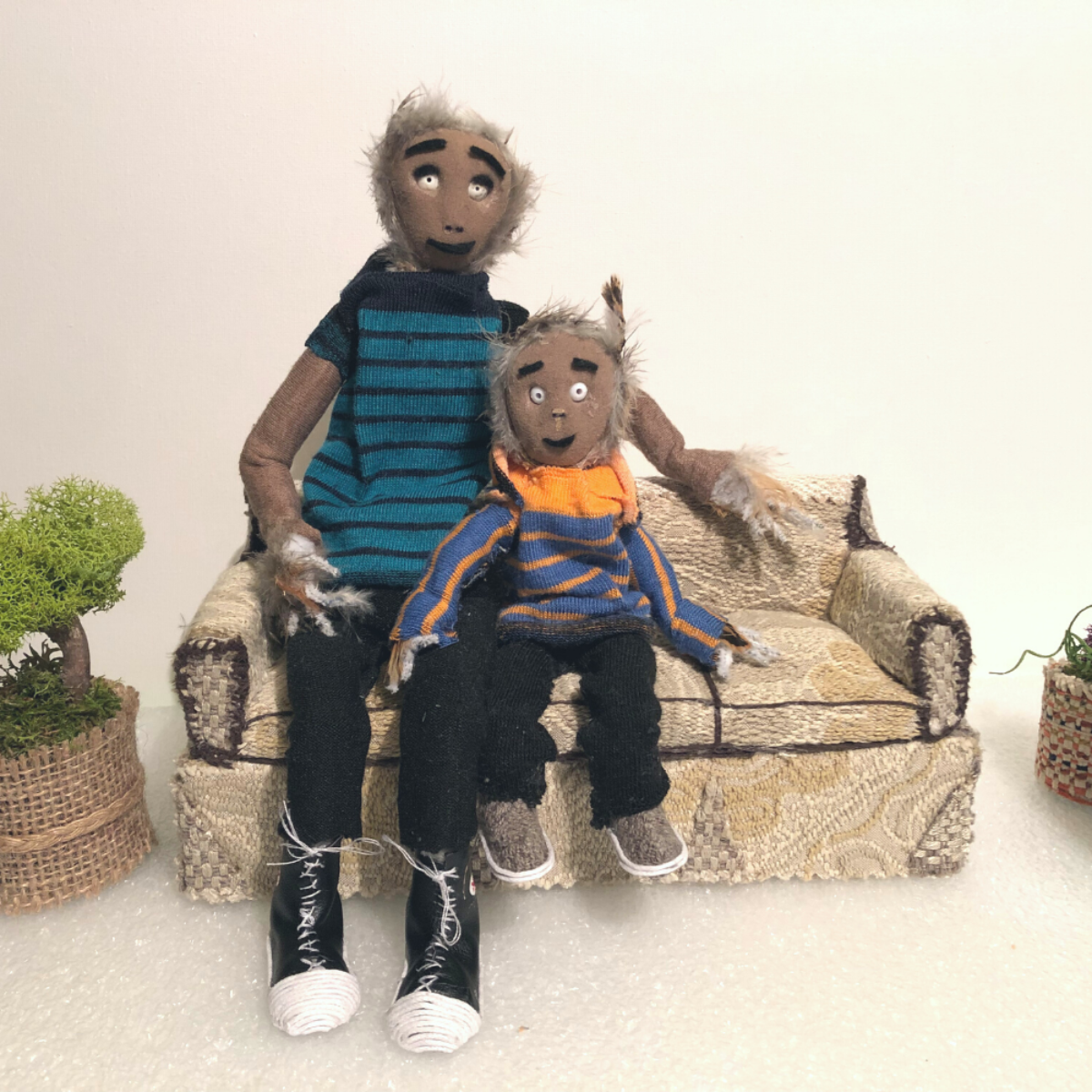 Two miniature stuffed figurines sit closely together on an old-fashioned miniature couch. The older one is wearing a blue shirt with black stripes, black pants, and white and black shoes. The younger one is wearing a blue and orange striped long-sleeve shirt, black pants, and grey and white shoes. On each end of the couch, there is a plant in a wicker basket.