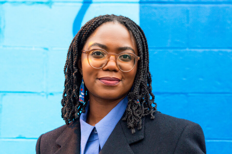 Chimwemwe smiles towards the camera as she stands against a blue painted brick wall. Her hair is black and styled in shoulder length braids. She is wearing round framed glasses, a blue collared shirt, and a dark grey blazer.