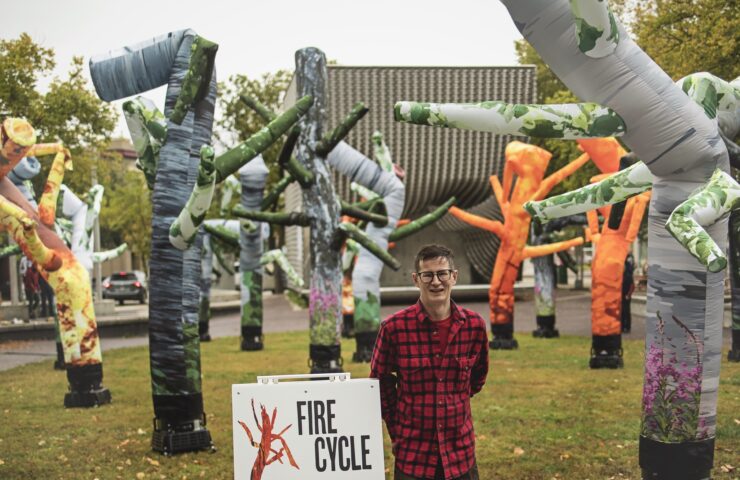 Artist Jonathan Green wearing a red flannel shirt, standing outside in front of 'Fire Cycle': an installation of various inflatable tubes designed as trees and flames.
