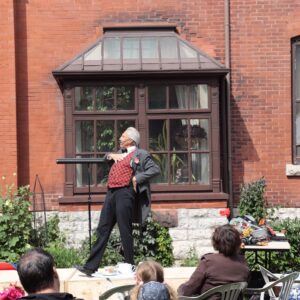 Grey-haired man dressed in a grey coat, red checkered vest, and black pants stands theatrically in front of a crowd on a stage. Behind him is a red brick building with large windows with brown shutters.