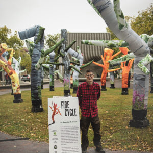 Artist in red plaid lumberjack shirt stands in front of inflatable tree artwork, beside sandwich board with title Fire Cycle.