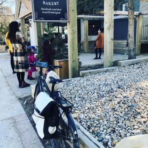A view from the sidewalk outside of a local bakery. There is a small line-up outside the bakery's entrance. A black bike is parked in front of a bed of various rocks.
