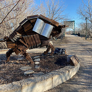 A sculpture rests on a raised platform along a sidewalk. The sculpture appears to be an abstract take on a buffalo, made out of rusted pieces of metal. In the background, the beginning of a bridge is visible and there are bare trees scattered about.