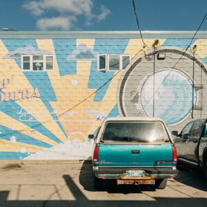 A shot from the back parking lot of a building shows two trucks, one teal and white and one black, parked in front of a mural on the backside of the building. The mural features blue and yellow beams emerging from the sun and a cloud.