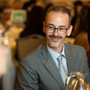 A man sitting at a table in a banquet-like setting. He is wearing grey suit, blue shirt, and silver tie, and sports glasses and a moustache.
