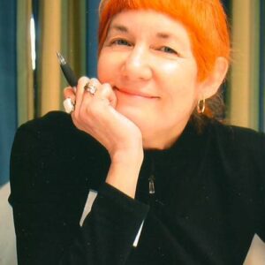 Red-haired woman wearing a black turtle neck rests her chin on her hand as she smiles at the camera.
